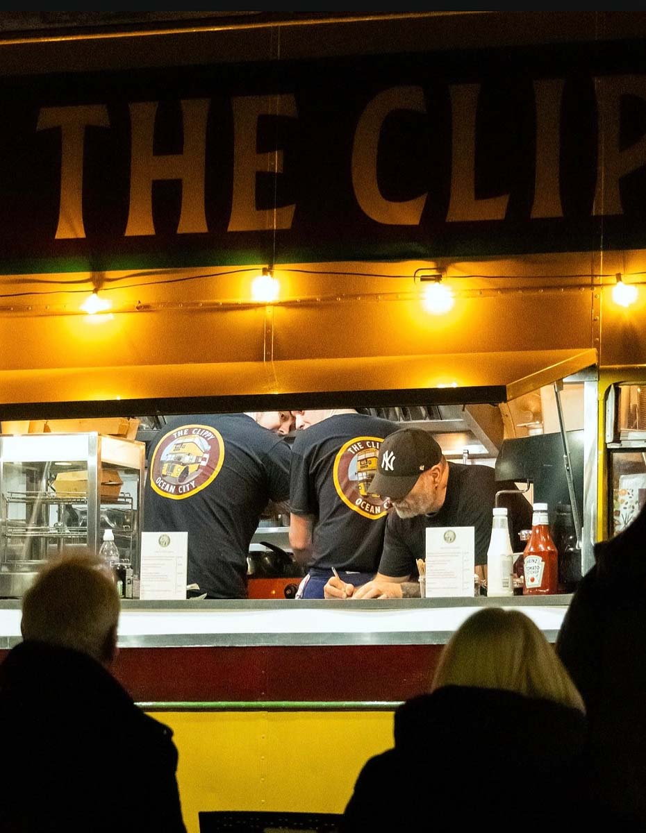 Discover The Clippy – A restored vintage bus on Plymouth Barbican, converted into a vibrant street food venue by Fletcher Andrews' father. The image shows the bus in the evening with a busy kitchen and a queue of eager customers ready to enjoy delicious street food.