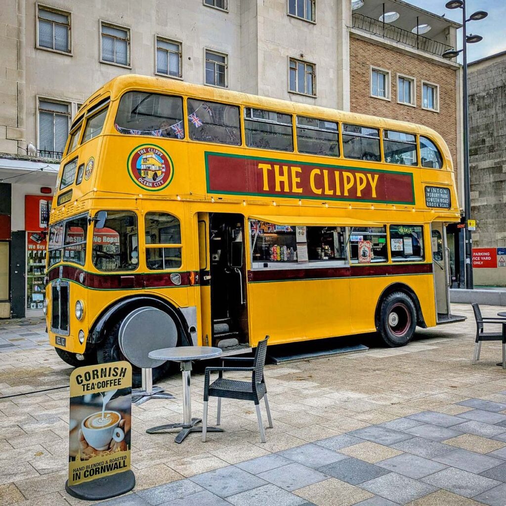 The Clippy, a bright yellow vintage double-decker bus converted into a street-food café, parked on New George Street in Plymouth, with outdoor tables and a sign advertising Cornish tea and coffee.